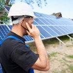 Back view of young engineer technician talking on cellphone standing in front of almost finished high exterior solar panel photo voltaic system in rural countryside on bright sunny summer day.