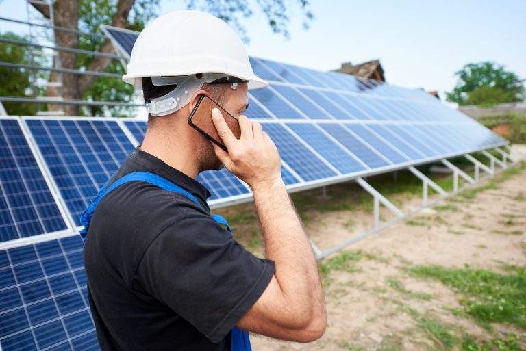Back view of young engineer technician talking on cellphone standing in front of almost finished high exterior solar panel photo voltaic system in rural countryside on bright sunny summer day.