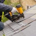 Unrecognizable roofer worker in special protective work wear and gloves, using air or pneumatic nail gun and installing asphalt or bitumen shingle on top of the new roof under construction residential building
