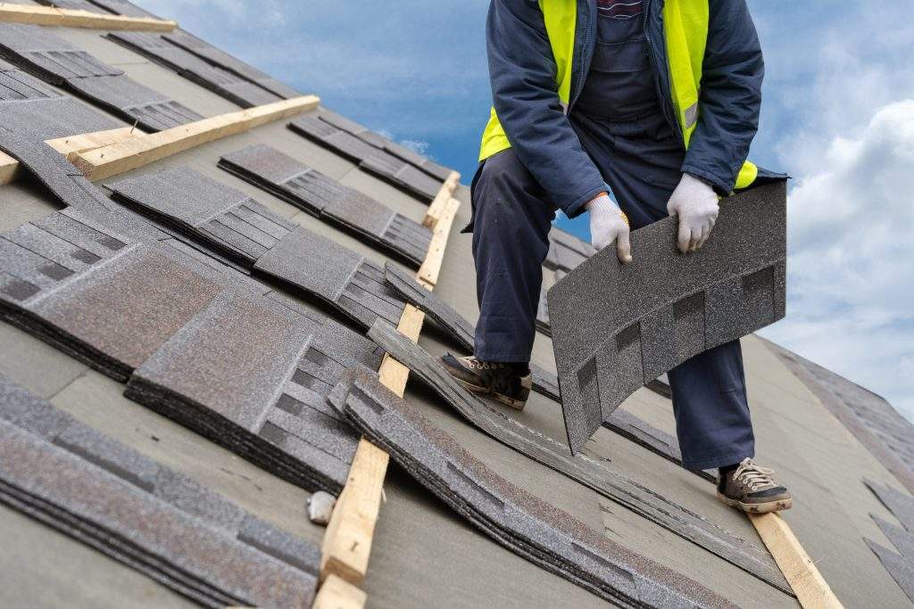 Qualified roofer worker in uniform work wear holding in hands and installing asphalt or bitumen tile on top of the roof under construction house against beautiful blue sky on background