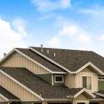 Panorama frame House exterior with view of the dark pitched roof against a cloudy blue sky. A mountain blanketed with snow can be seen behind the home.
