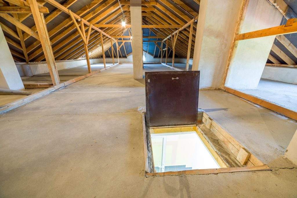Attic of a building with wooden beams of a roof structure and a fire exit door in floor.